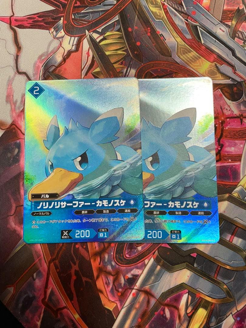 Two holographic trading cards of the ノリノリサーファーカモノスケ promo shown overlapping on a patterned surface.