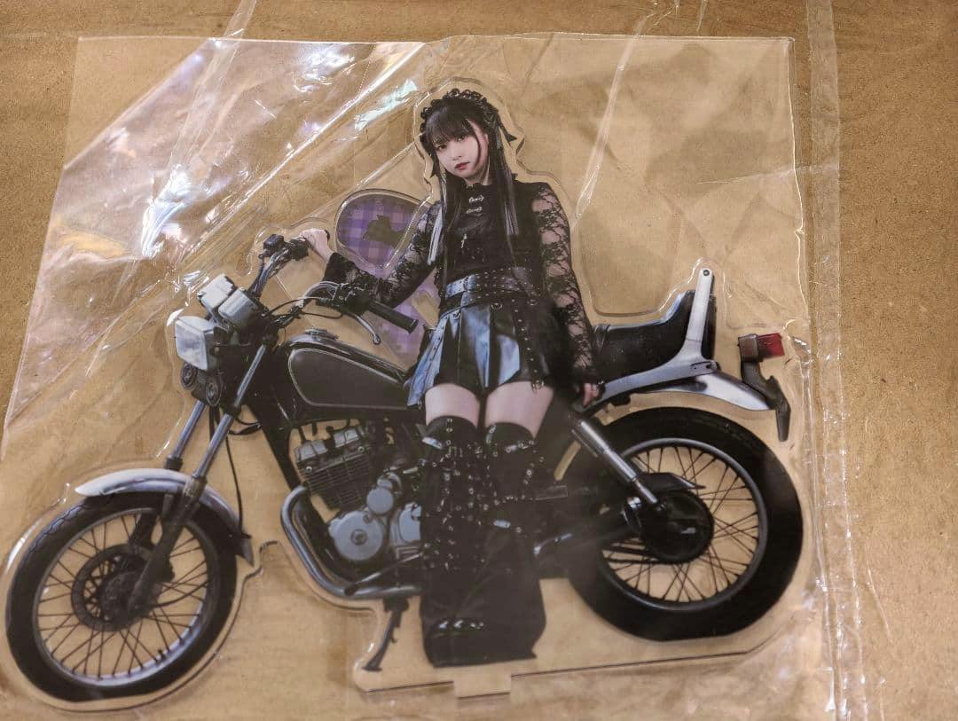 Die-cut acrylic stand showing a dressed figure sitting on a motorcycle, inside a clear plastic sleeve on cardboard.