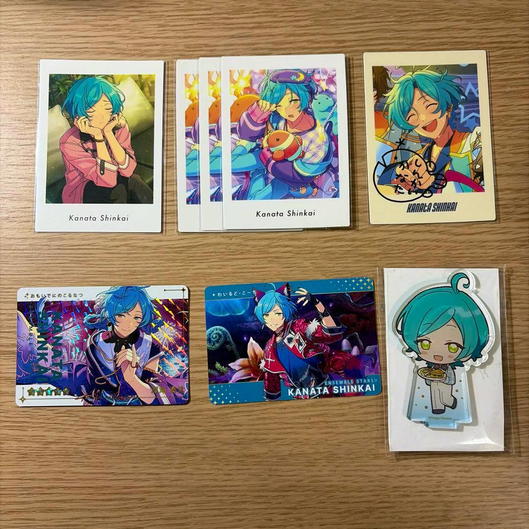 Photo showing multiple Kanata Shinkai trading cards and a packaged acrylic stand arranged on a wooden surface.