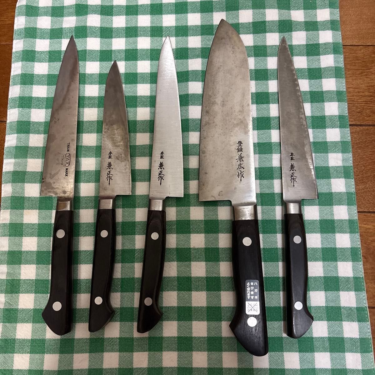 Five steel chef knives laid out side-by-side on a green-and-white checkered cloth.