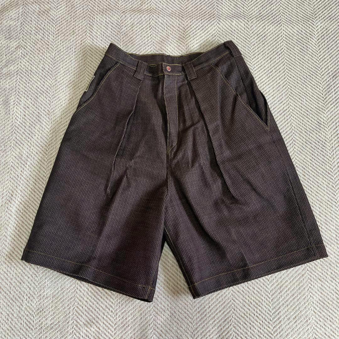 Front view of dark woven denim men's shorts laid flat on a patterned surface, showing the waistband, front pockets, pleats, and hem.