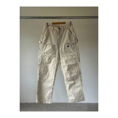 Front view of KAPITAL Light Canvas Lumbar Pants in natural white hanging on a wooden hanger against a plain wall.