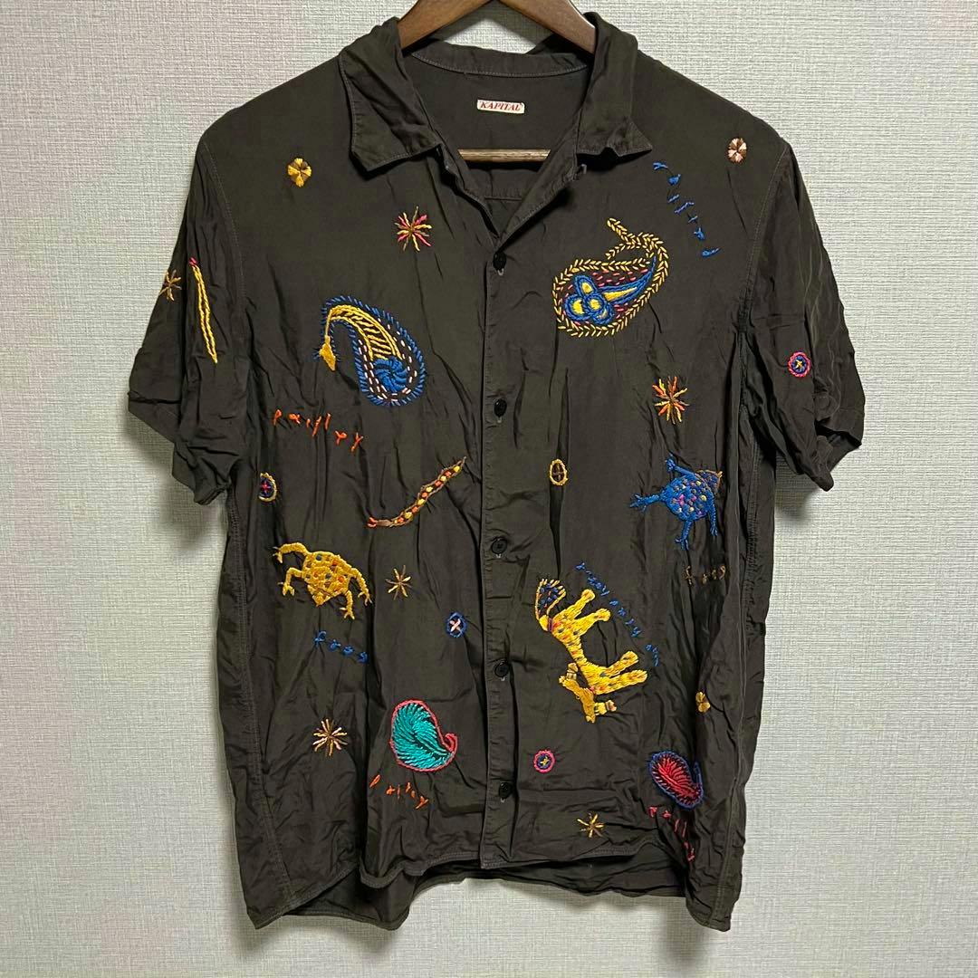 Front view of a KAPITAL men's rayon short-sleeve shirt with colorful embroidered motifs hanging on a wooden hanger.