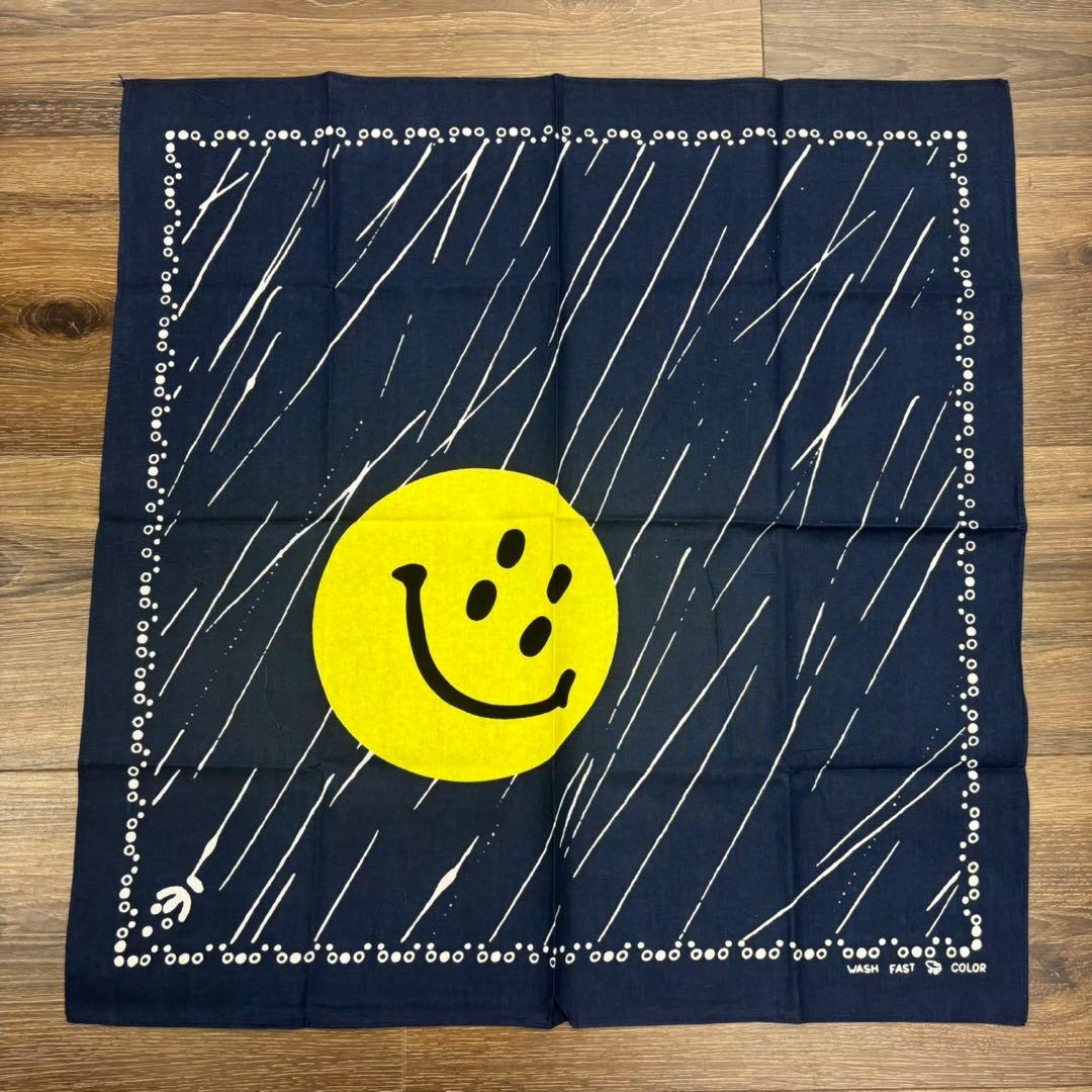 A navy cotton bandana laid flat showing a large yellow smiley face at center with white diagonal streaks and a dotted border on a wooden background.