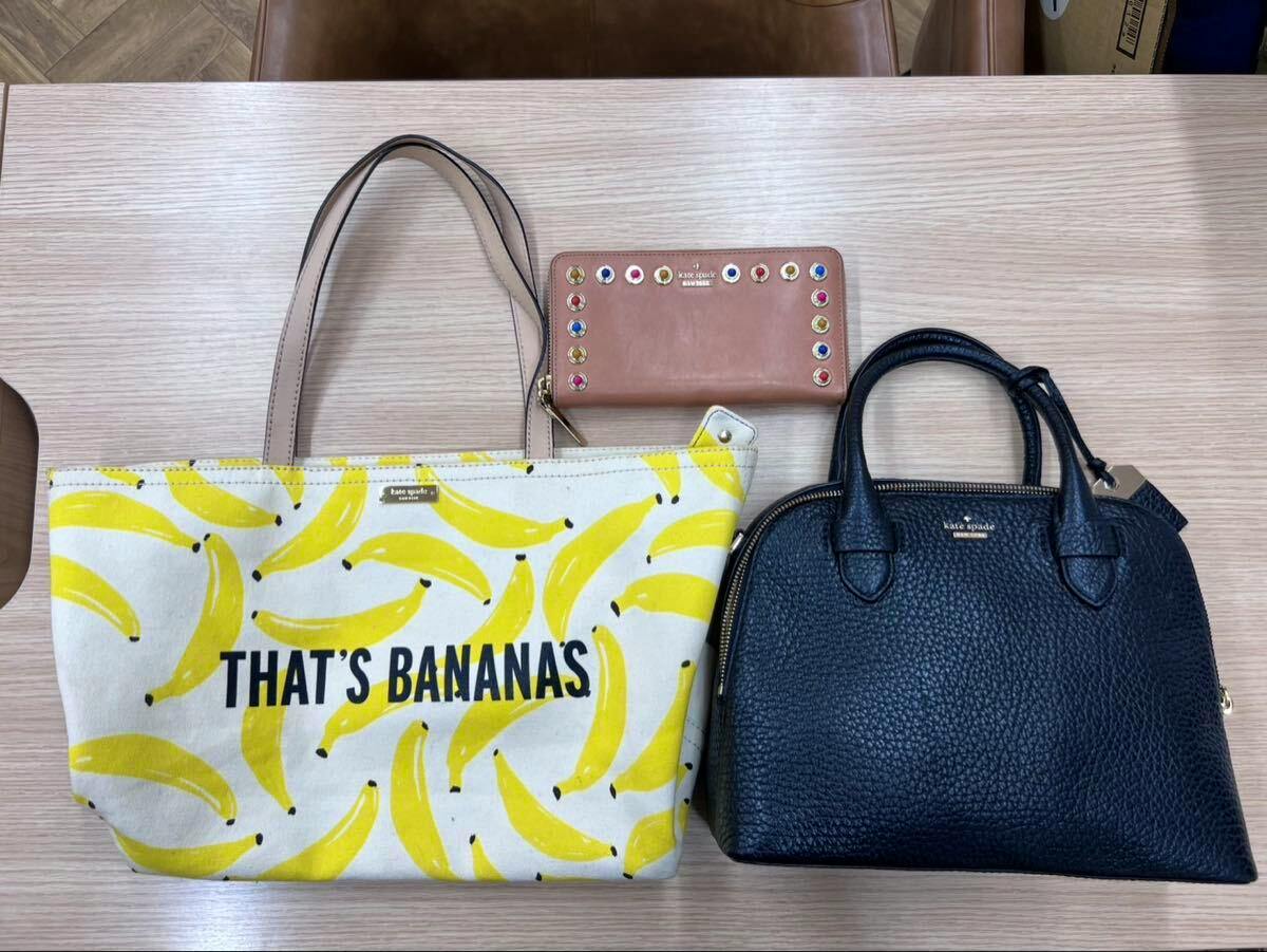 Three Kate Spade items on a table: a banana-pattern canvas tote with "THAT'S BANANAS" text, a black leather handbag, and a pink long wallet.