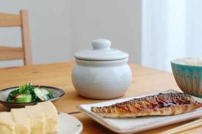A small white ceramic pot with a lid sitting on a dining table among plates of food.