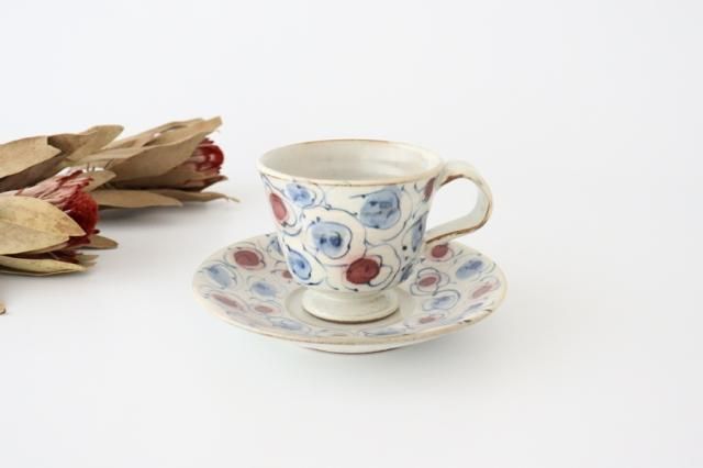 Goblet-shaped ceramic coffee cup and matching saucer with blue and red circular floral motif on a white surface with dried flowers to the left.