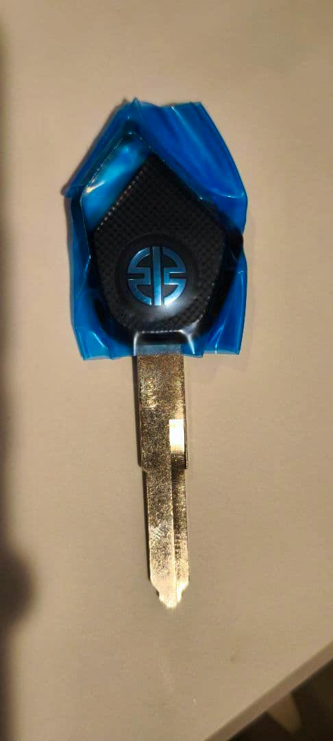 Photo of the Kawasaki spare blank motorcycle key with a black-and-blue plastic head wrapped in blue film and an uncut metal blade on a pale background.