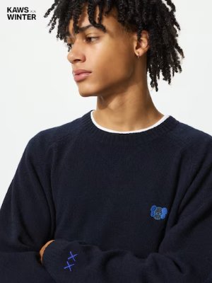 Model wearing a navy crewneck sweater with a small blue embroidered KAWS character on the chest.