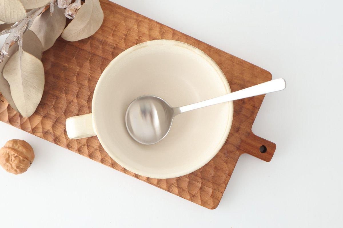 A metal soup spoon resting inside a ceramic bowl on a wooden cutting board.