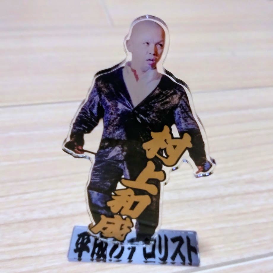 Photograph of the 村上和成アクリルスタンド acrylic stand showing the cutout figure of a man in a suit mounted on a rectangular base with gold Japanese text.