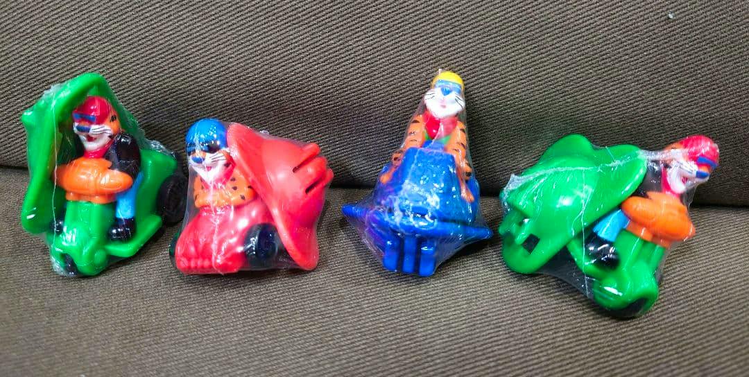 Four small plastic Kellogg promotional figures in colored vehicles, wrapped in clear plastic and arranged on a brown fabric surface.