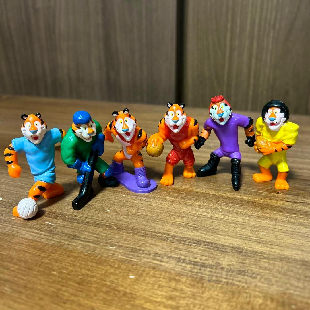 Six small Tony the Tiger plastic figures in different colored sports outfits on a wooden surface.