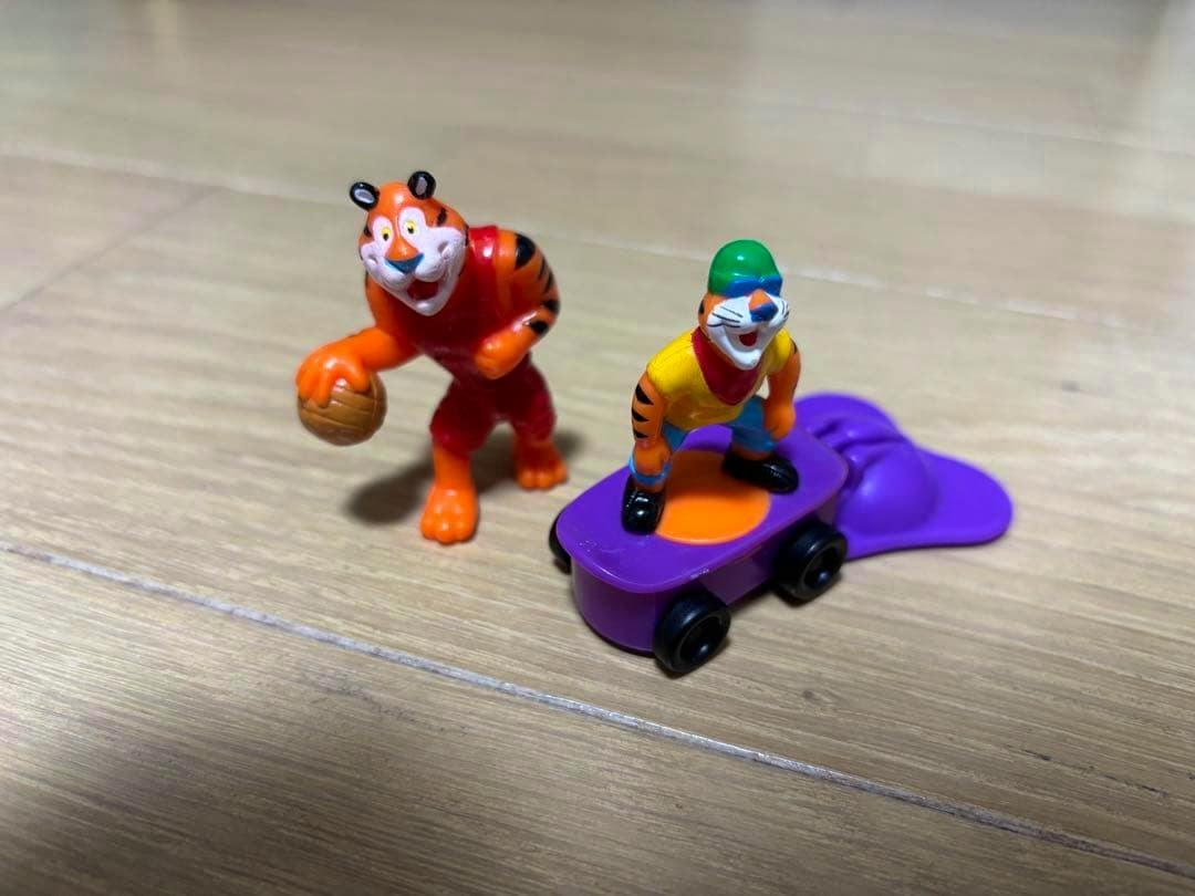 Two small Kellogg tiger plastic figures on a wooden surface, one holding a basketball and one standing on a purple skateboard-like roller.