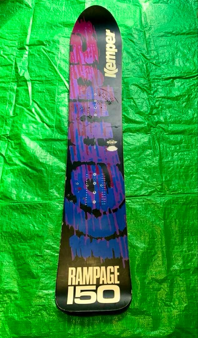Top view of a Kemper Rampage 150 snowboard showing purple and black graphics with 'RAMPAGE 150' text on a green background.