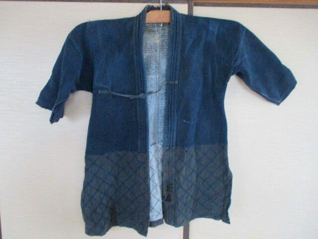 Front view of a blue indigo cotton kendo jacket (top only) hanging on a wooden hanger against a white wall.