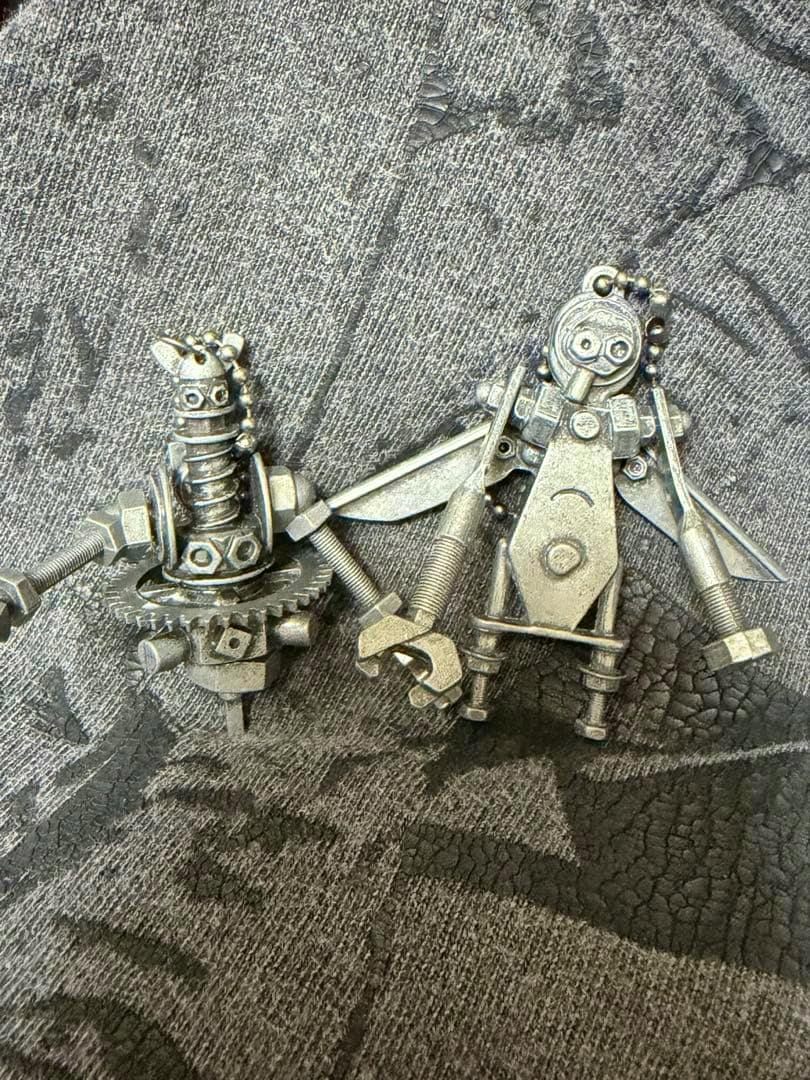 Two silver-colored metal bag charms/keyholders shaped like small robots placed on textured fabric.