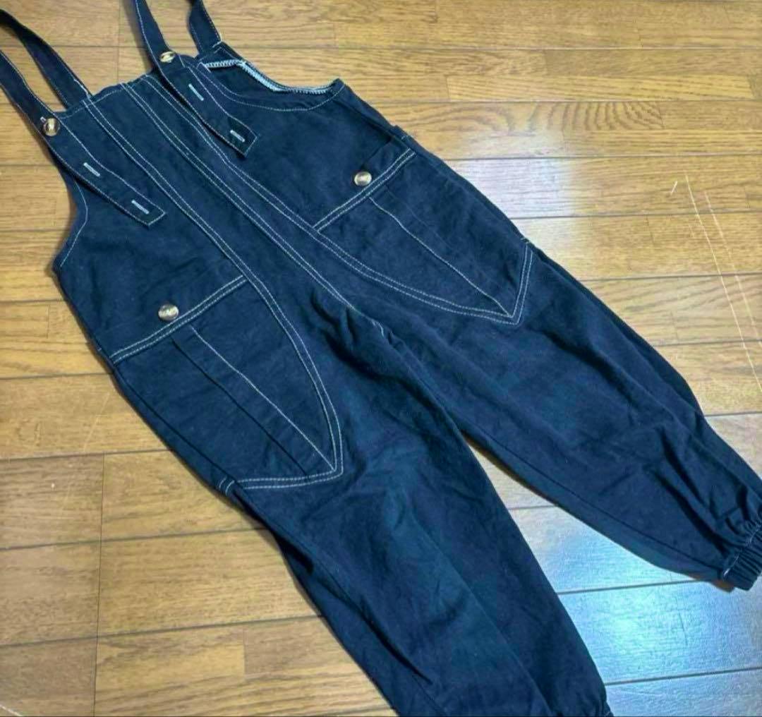 Flat-lay photo of black kids' cotton overalls with front pockets, contrast stitching, and shoulder straps on a wooden floor.