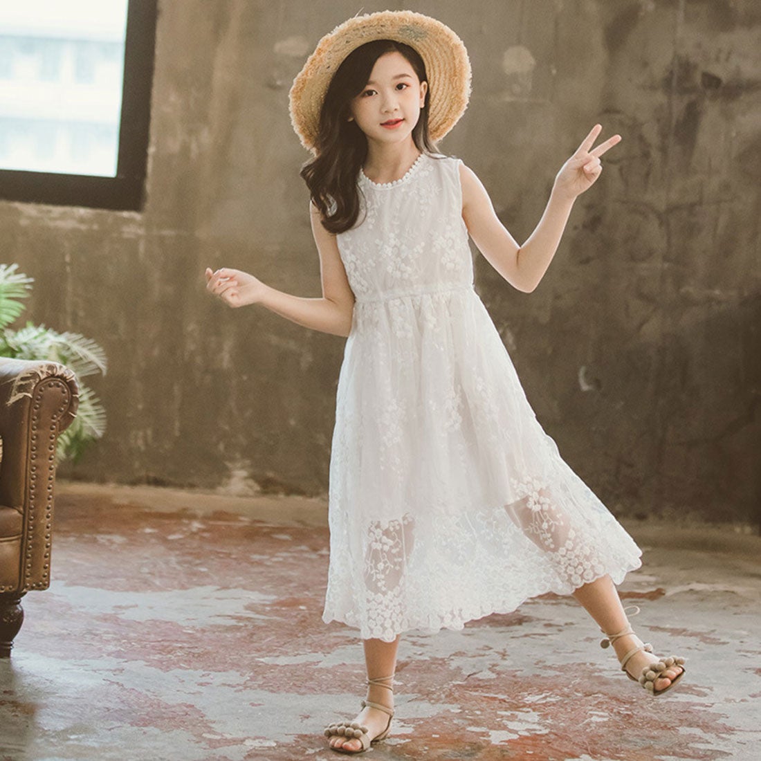 Child model wearing a white kids' sleeveless lace dress and a straw hat standing indoors.