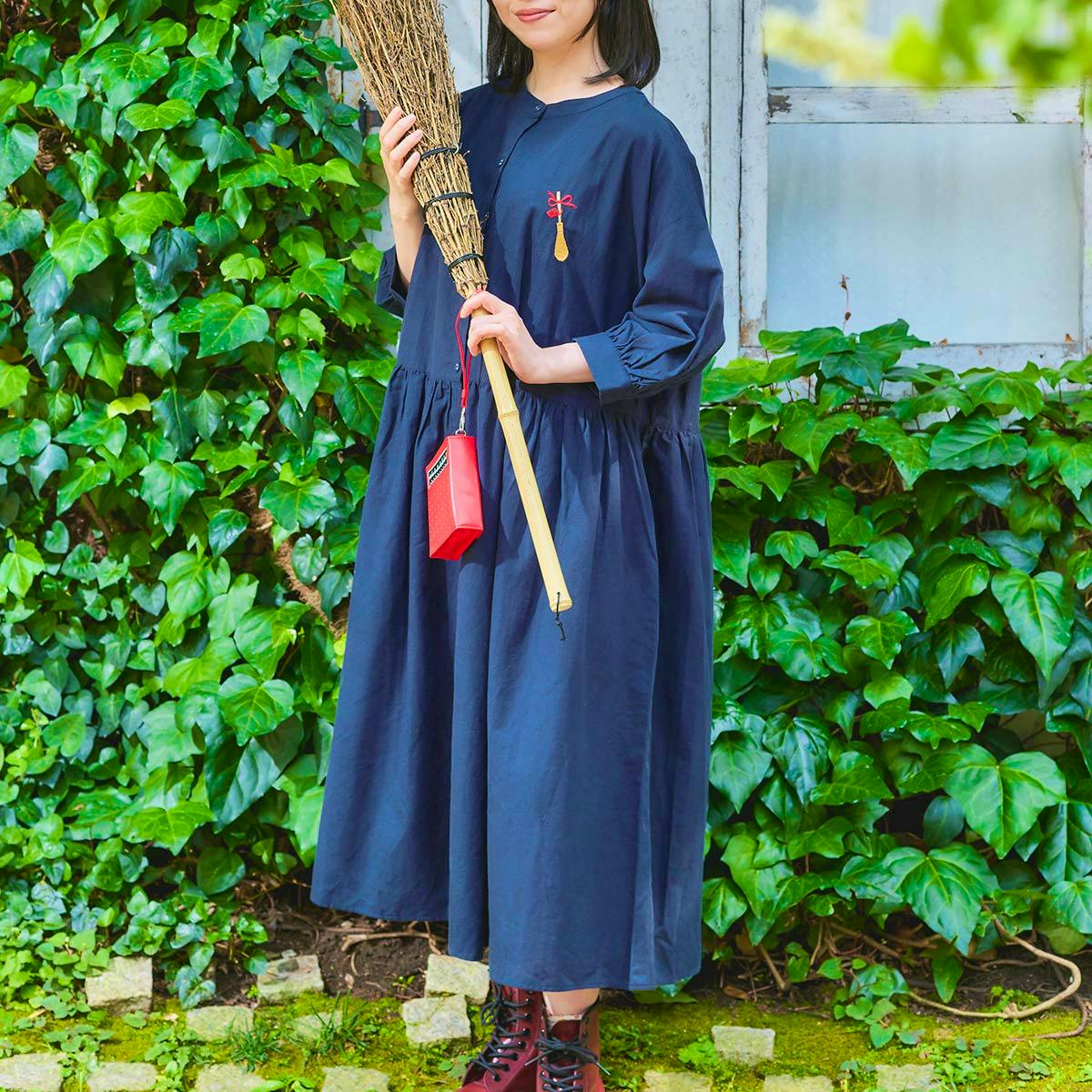 A model wearing a navy cotton 2-way shirt dress stands outdoors holding a broom in front of green ivy.