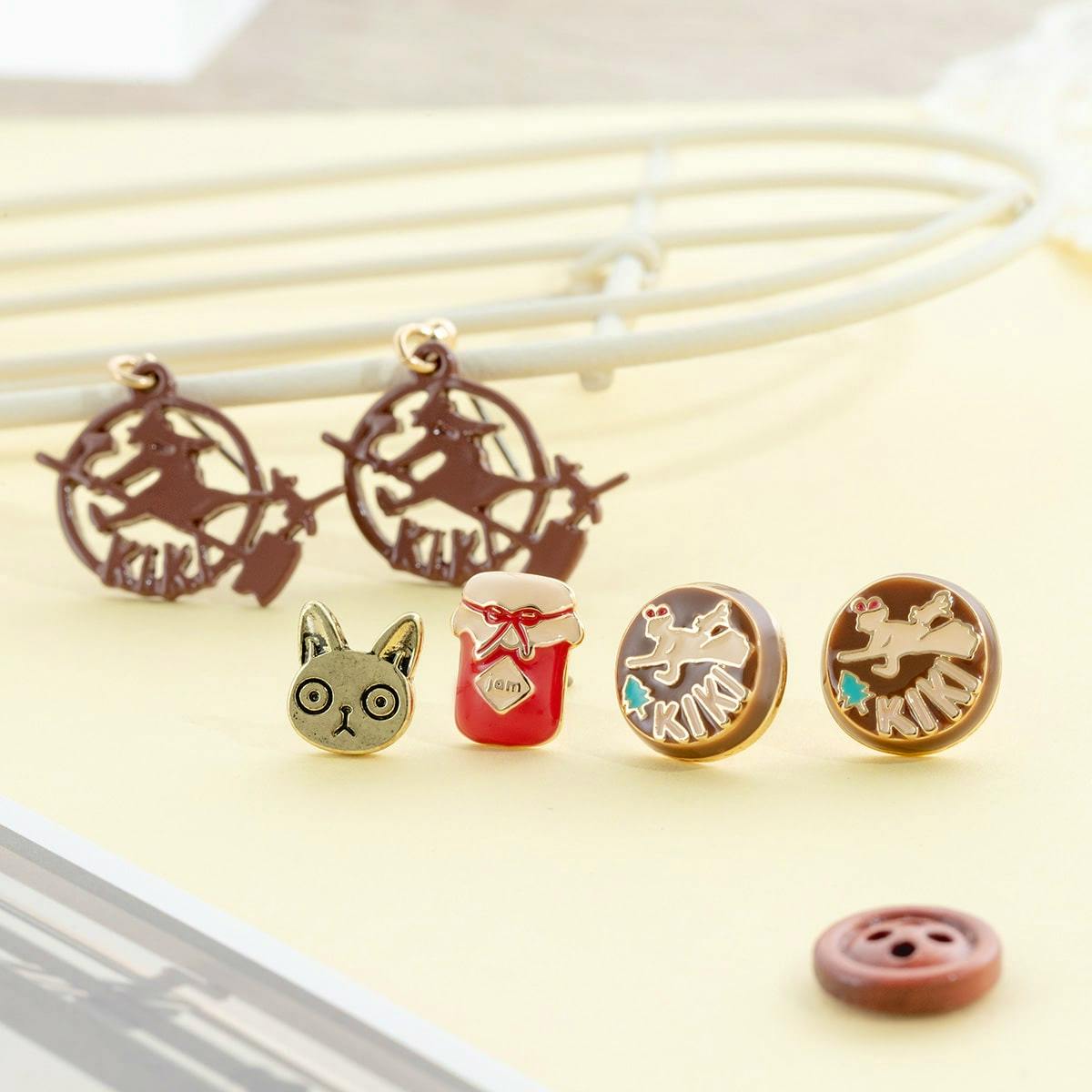 Six Kiki's Delivery Service themed non-precious metal earrings arranged on a pale surface, showing a cat face, a jam jar, two round "KIKI" studs, and two witch-on-broom dangle charms.