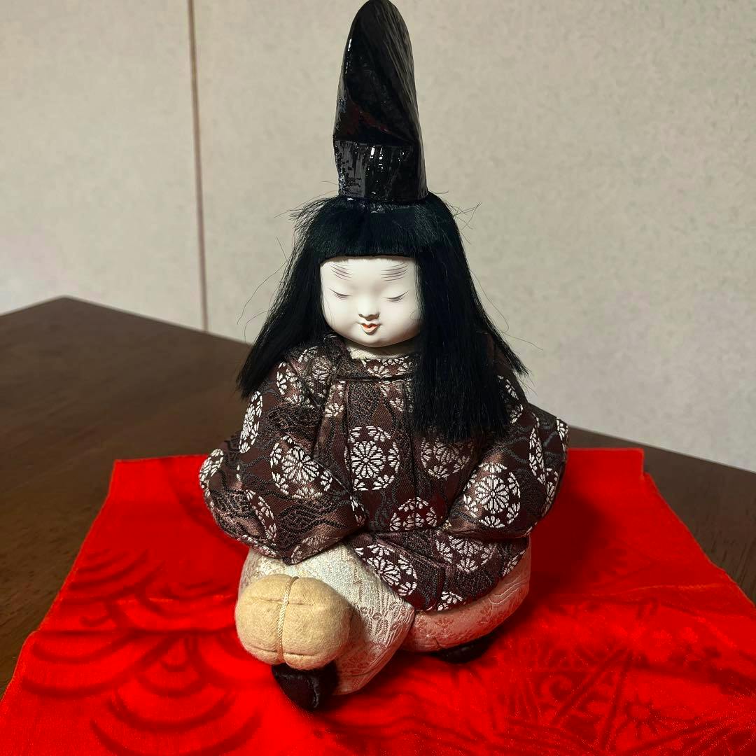 Front view of a wooden kigome doll of a child in a patterned kimono seated on a red cloth with a small ball at its feet.