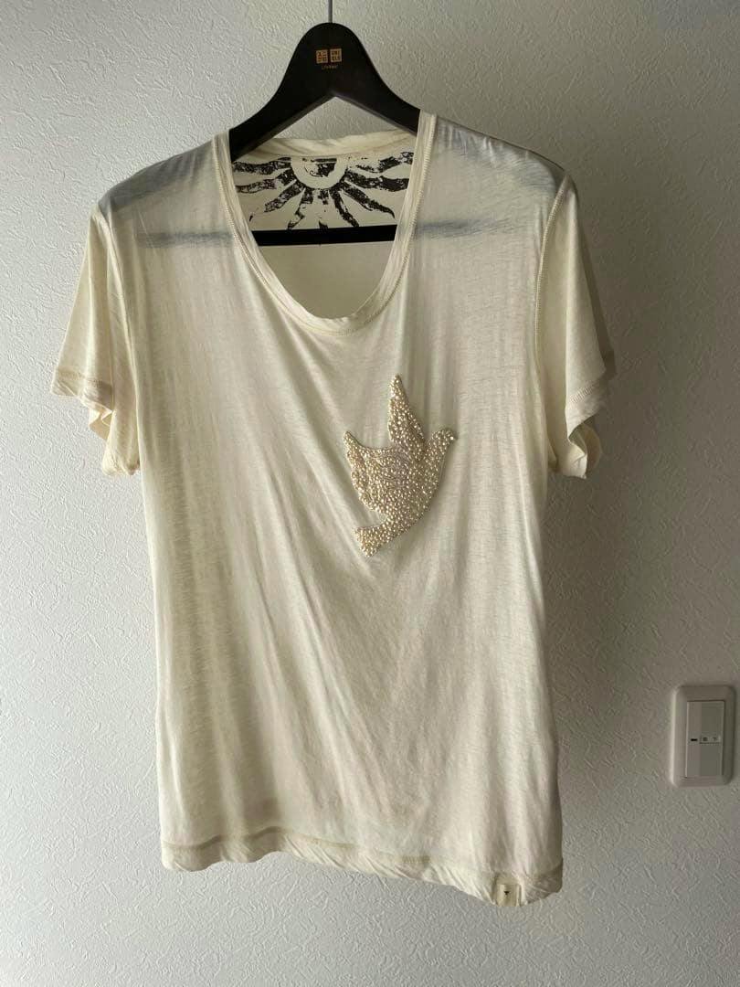 Off-white cotton T-shirt with a pearl-decorated bird motif hanging on a hanger against a wall.