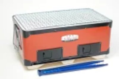 Red rectangular tabletop charcoal barbecue grill with a metal cooking grate and a pair of blue tongs in front.