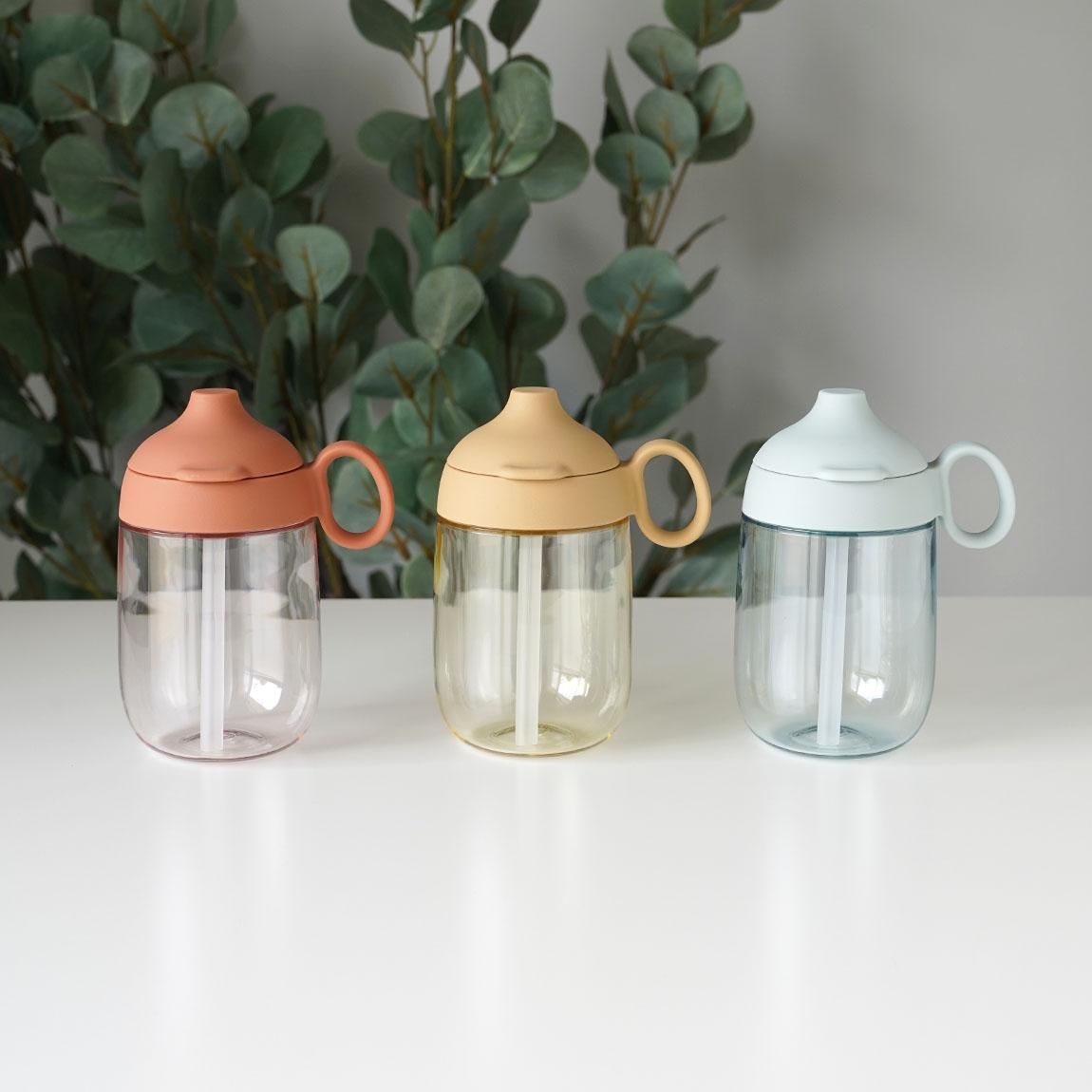 Three Kinto BONBO 260ml plastic straw mugs in three different colors arranged on a white surface with foliage in the background.