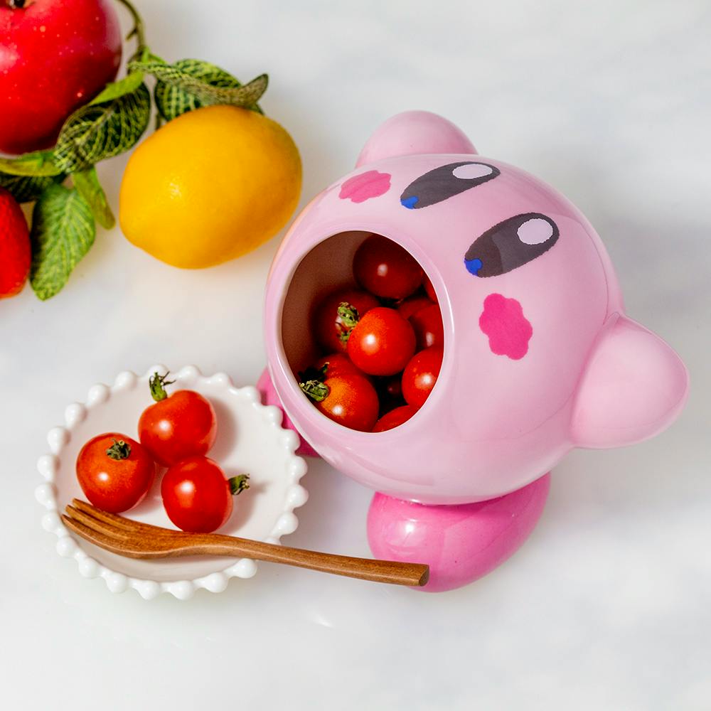 Pink Kirby-shaped ceramic salad bowl with an open mouth filled with cherry tomatoes, shown alongside a small white dish, wooden fork, and fruit props.