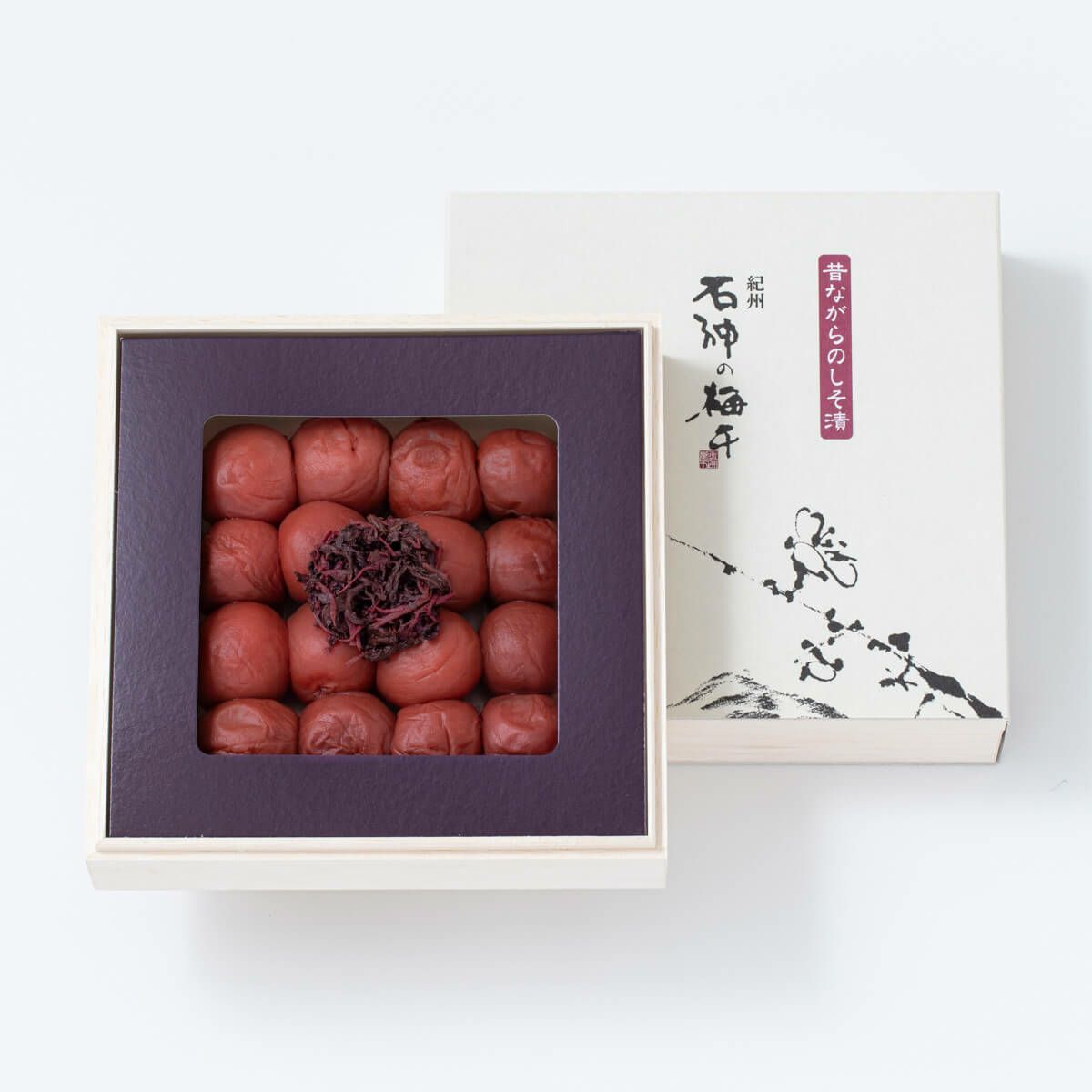 An open square box of shiso-pickled preserved plums arranged in a grid with a pile of pickled shiso in the center and the decorative box lid beside it.