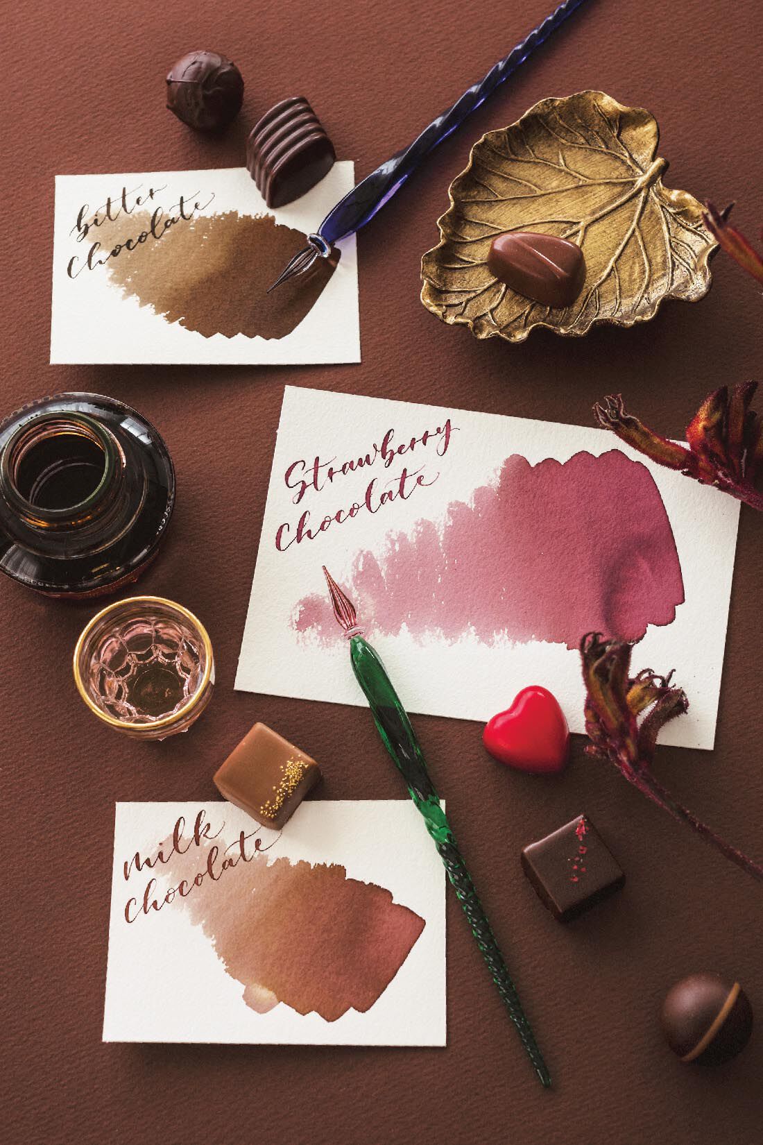 A tabletop arrangement showing fountain-pen ink swatches labeled "Strawberry chocolate" and "milk chocolate", an open ink bottle, glass dip pens, and chocolate pieces on a brown background.