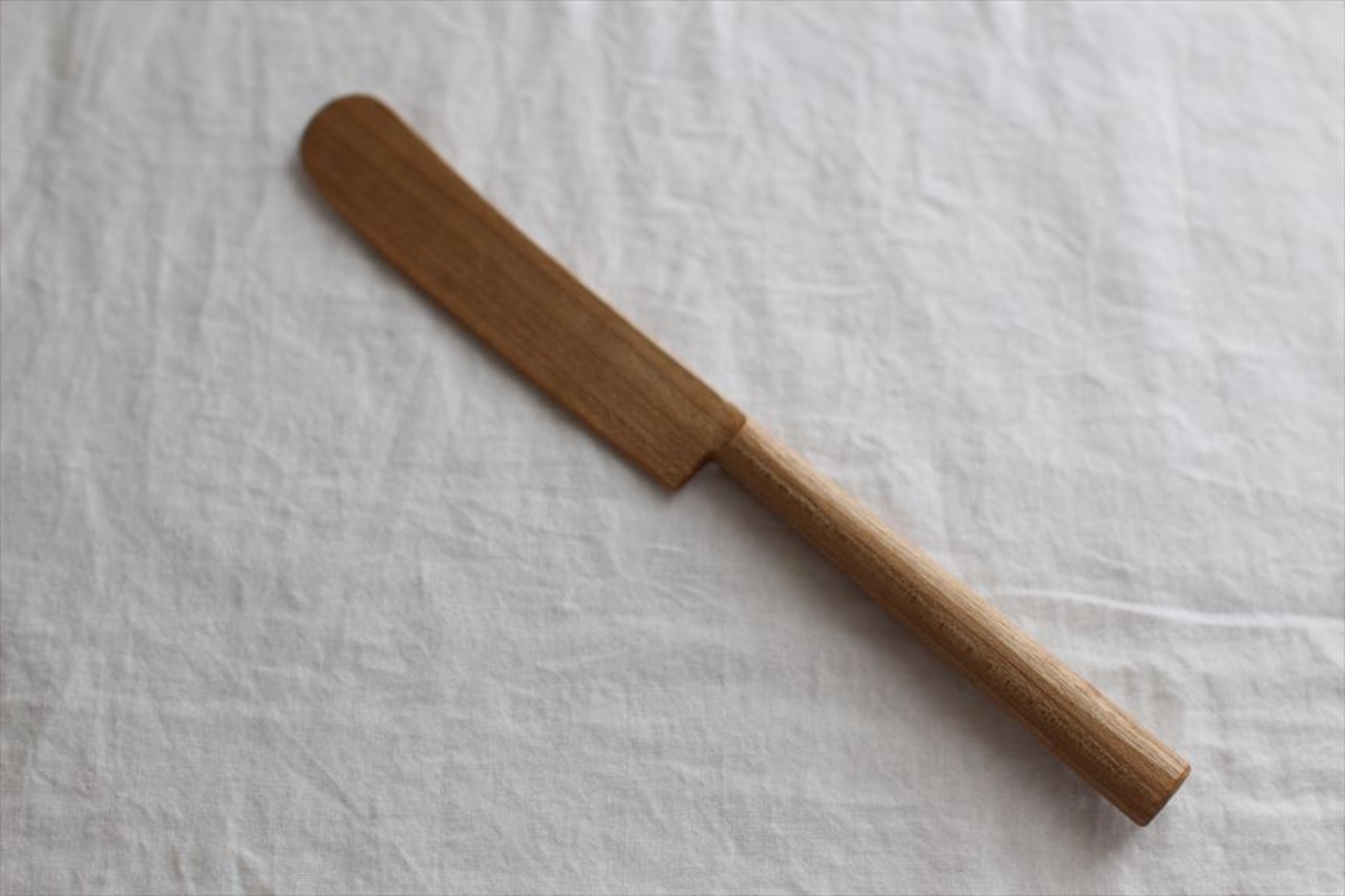 Wooden butter knife with a rounded blade and cylindrical handle placed diagonally on a white cloth background.