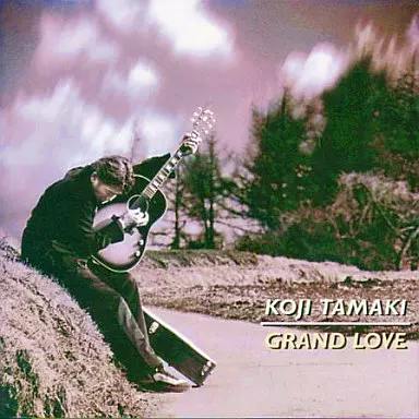 CD cover art for 玉置浩二 / GRAND LOVE showing a person seated on a roadside playing a guitar with trees and sky in the background and the text "KOJI TAMAKI GRAND LOVE".