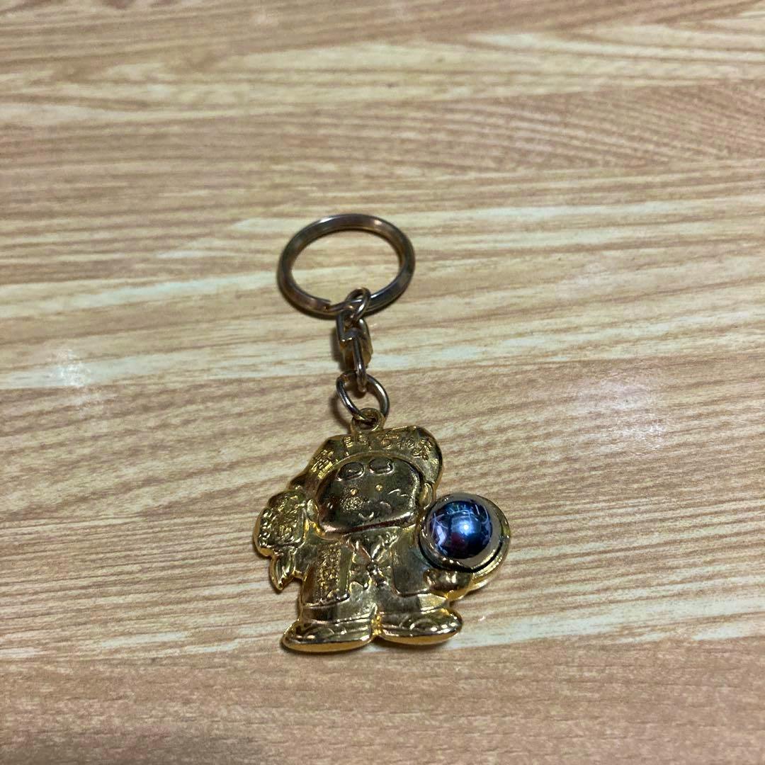 Gold-colored zinc alloy bag charm keychain shaped like a character holding a small sphere, photographed on a wood surface.