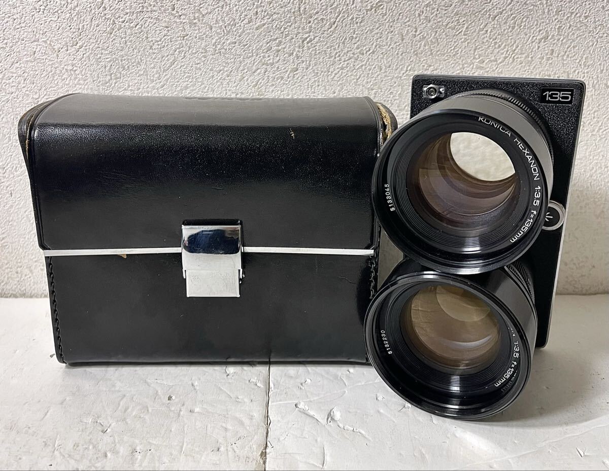 Twin-lens reflex camera showing a KONICA HEXANON 135mm lens mounted, with a black leather case beside it.