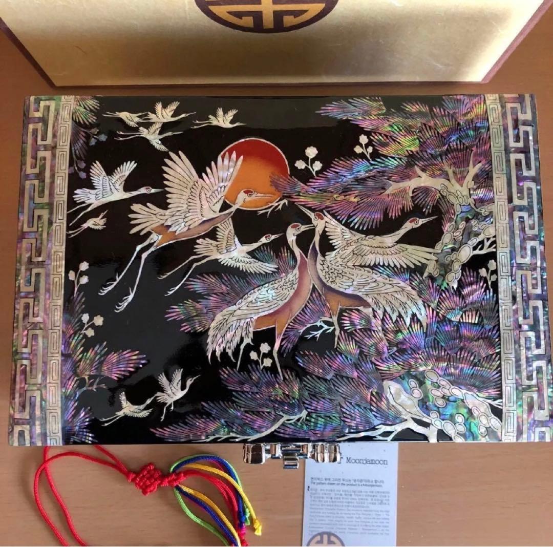 Top view of a wooden Korean mother-of-pearl inlay jewelry box with crane design, metal latch, multicolored tassel, and an information card.