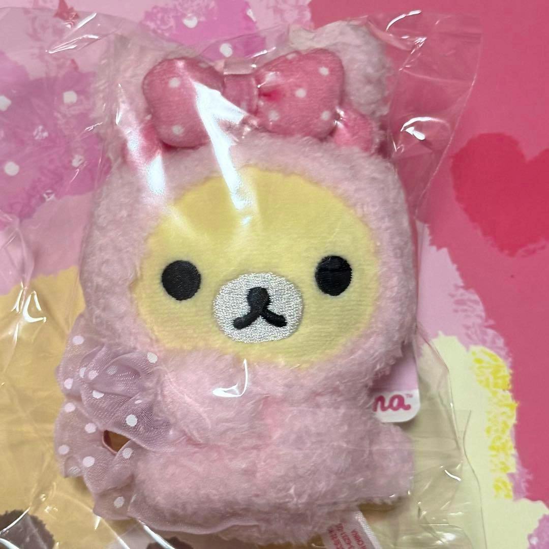 A pink Korilakkuma plush wearing a bunny hood shown inside clear plastic packaging on a pink background.