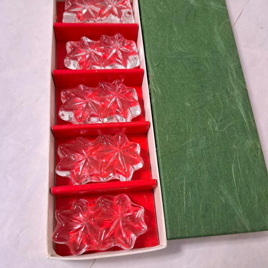 Five clear crystal chopstick rests arranged in a red-lined box beside a green textured lid.