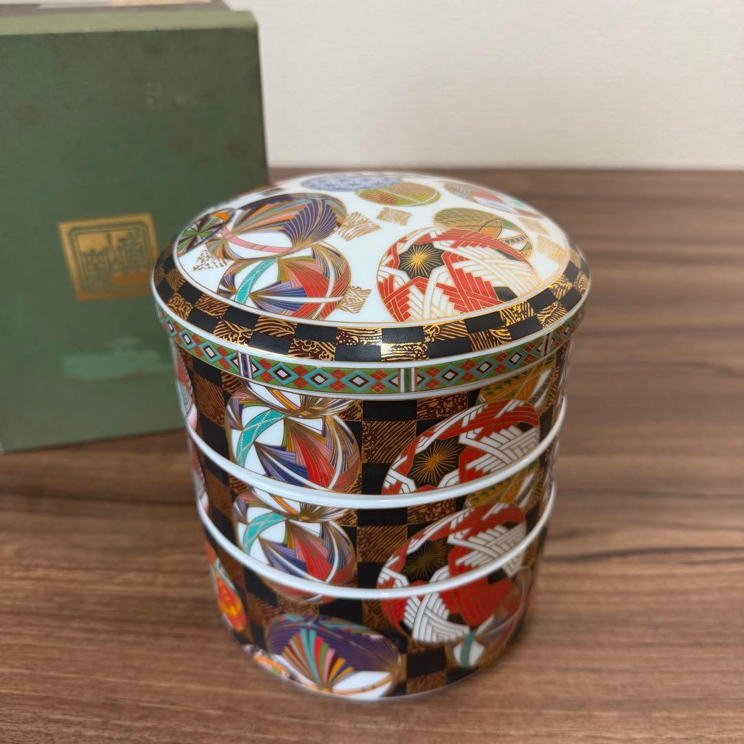 Three-tiered ceramic stacking box with lid, decorated with colorful temari ball motifs, gold highlights, and a checked pattern, shown on a wooden surface.