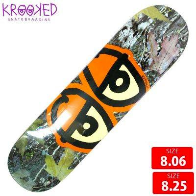 Krooked Team Eyes camo skateboard deck shown flat with a large orange eye graphic and visible size labels.