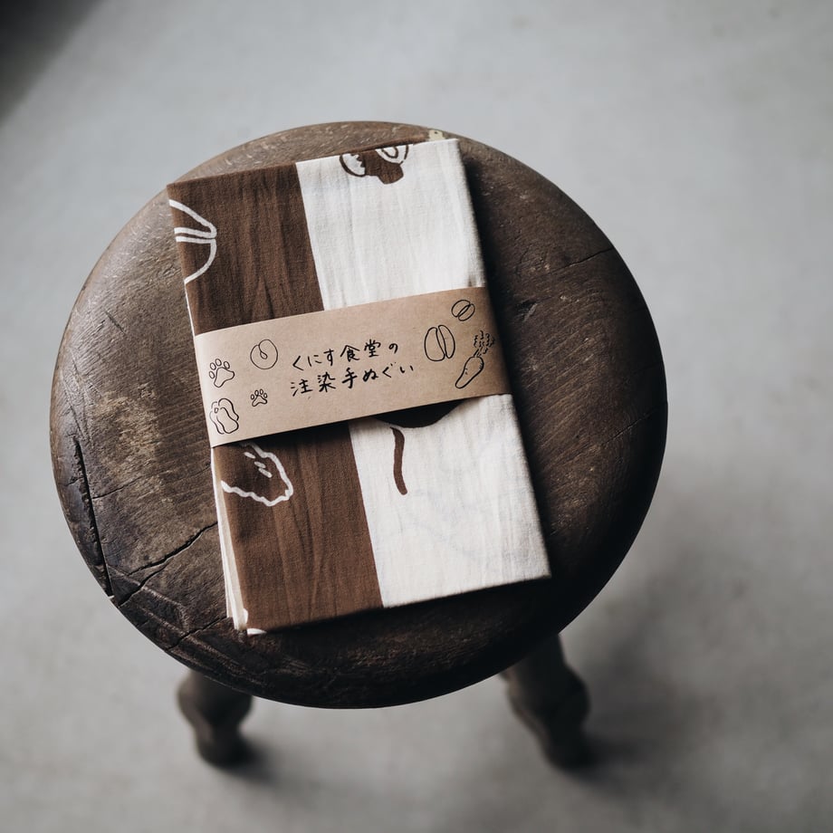 A folded cotton tenugui hand towel with a paper band label placed on a round wooden stool.