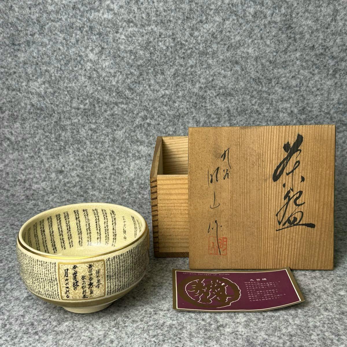 A Kutani ceramic matcha tea bowl decorated with dense calligraphic text and gold trim, shown with its wooden storage box and an information card.