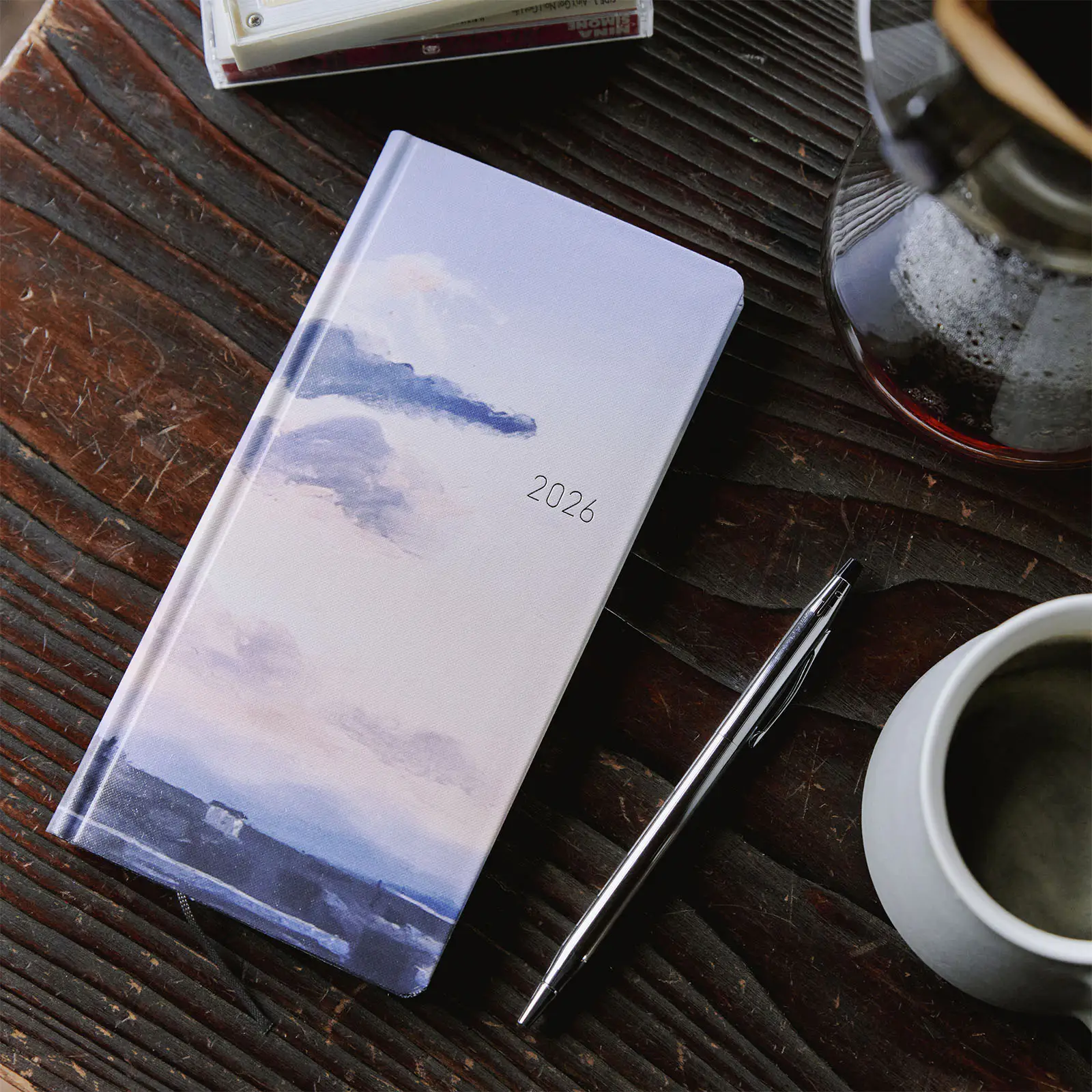 Kyohei Sakaguchi hardcover weekly planner with a dawn landscape cover and “2026” on the front, placed on a wooden table next to a pen and a cup.