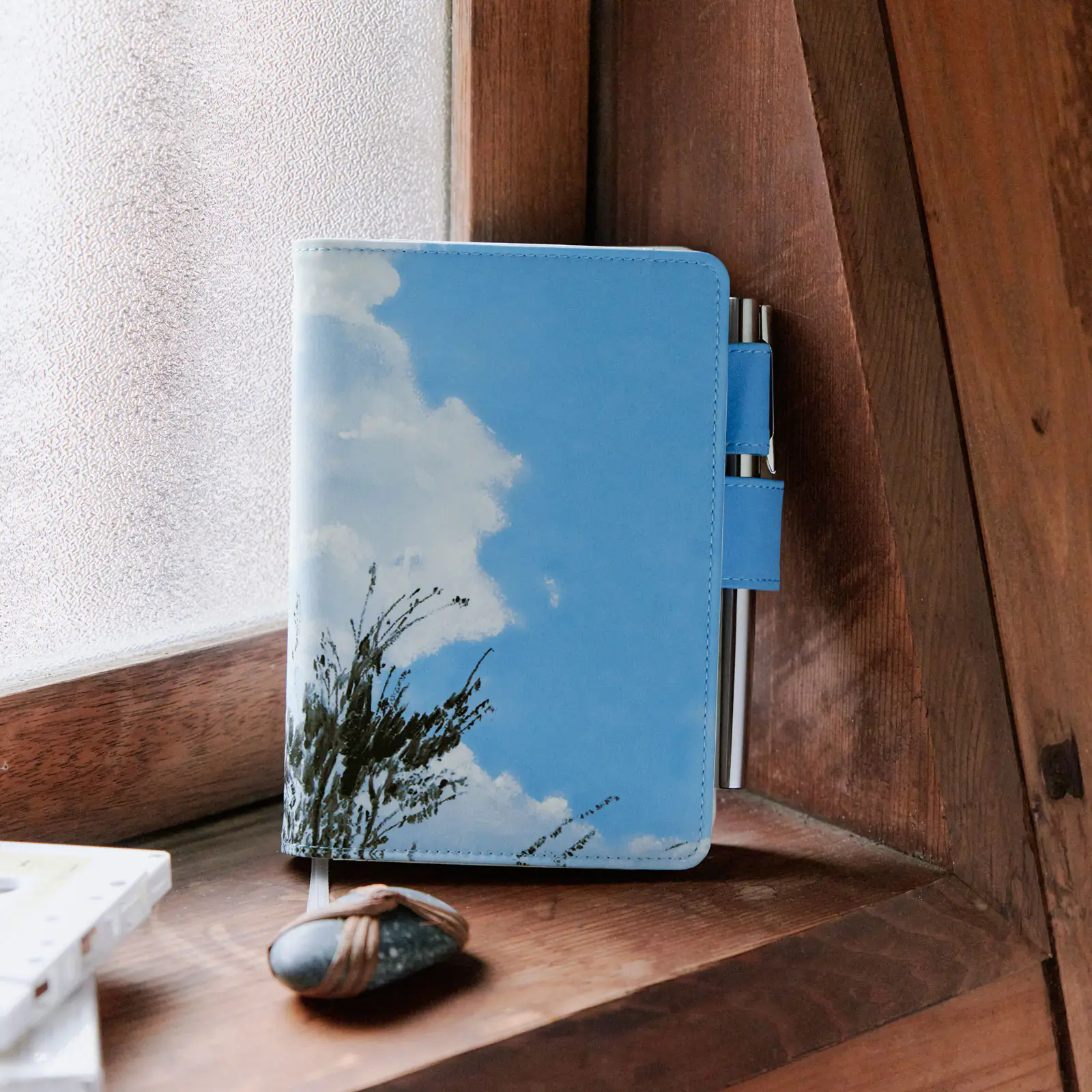 A blue sky-and-cloud A6 polyester book cover standing on a wooden windowsill with a pen in its pen loop.