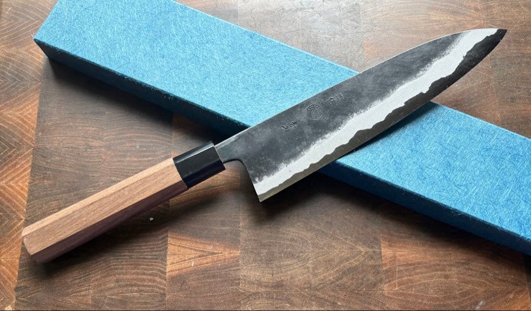 A 210mm gyuto chef knife with a dark hammered blade and wooden handle placed on a blue box over a wood surface.