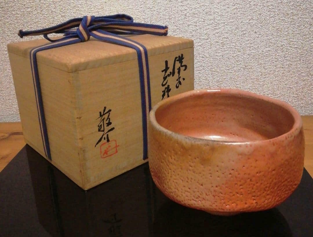 A Shino-style tea bowl (chawan) in pinkish glaze displayed beside its signed wooden storage box tied with a ribbon.
