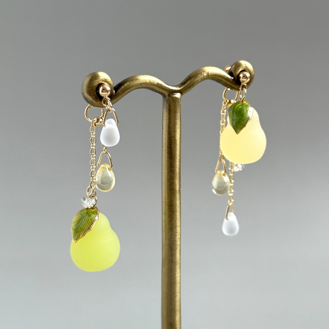 Asymmetrical La France bonbon earrings in yellow-green with leaf charms and dangling beads displayed on a metal stand.