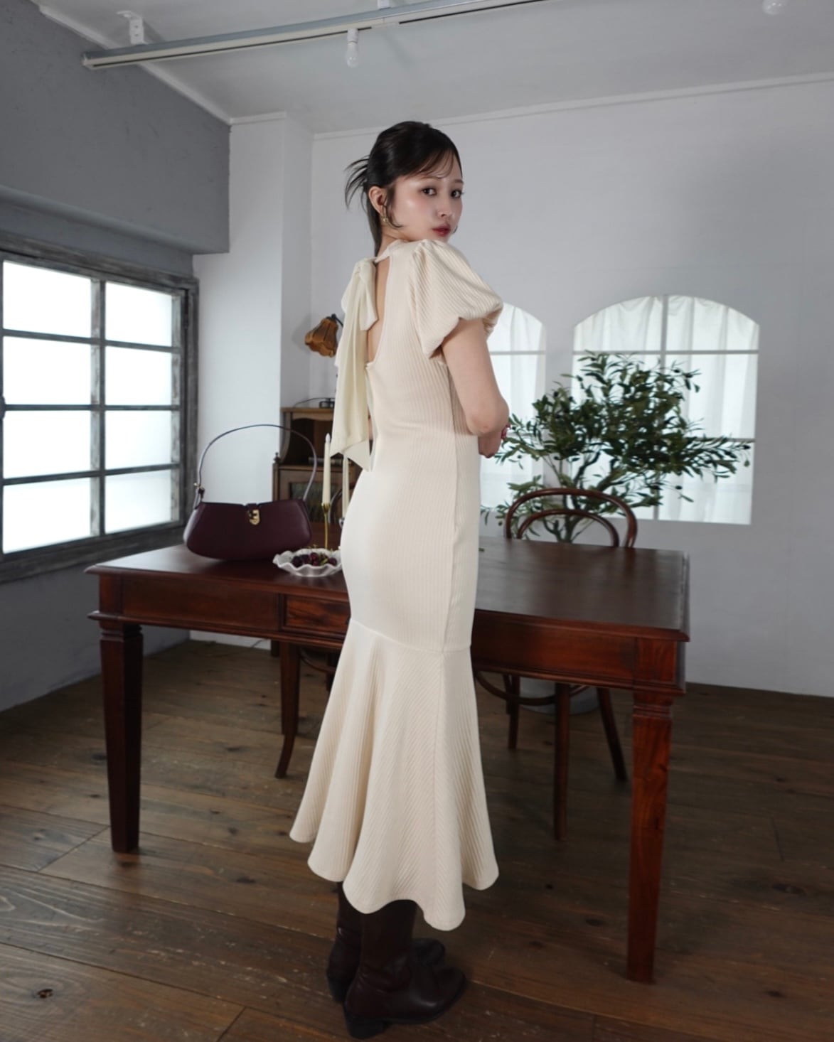 Woman wearing a cream mermaid-style women's polyester dress, shown from the back standing beside a wooden table in a room.