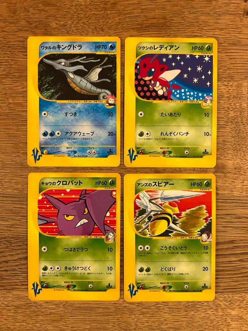 Four Japanese Pokémon VS trading cards showing ワタルのキングドラ, ツクシのレディアン, キョウのクロバット, and アンズのスピア on a wooden surface.